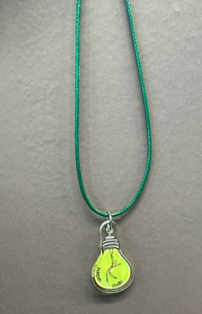 A green cord necklace featuring a lightbulb-shaped pendant with a glowing neon yellow-green interior, a black dancing figure, and "Modern Dance Studio" text displayed on a gray background