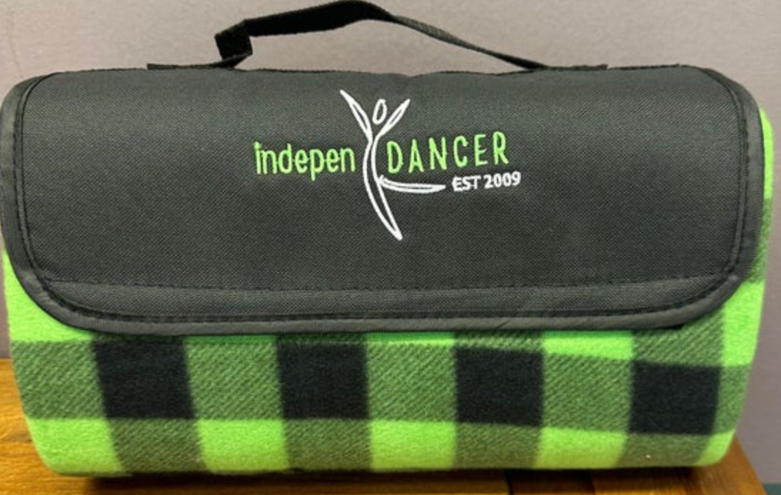 A folded picnic blanket featuring a black carrying flap with the "indepen DANCER EST 2009" logo in white and green, revealing a vibrant lime green and black buffalo plaid pattern