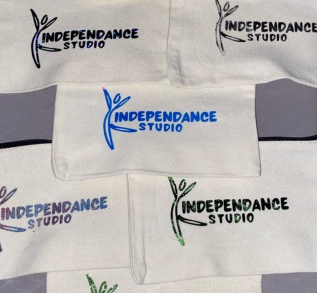 Multiple white textile pouches showcasing the INDEPENDANCE STUDIO logo with a dancing figure design in varied colors including prominent blue black iridescent and green glitter finishes