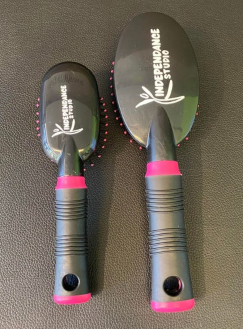Product photo of two black hairbrushes, one small and one large, with vibrant pink bristles and handle accents, featuring the white INDEPENDANCE STUDIO text and dancing figure logo, all on a dark textured surface