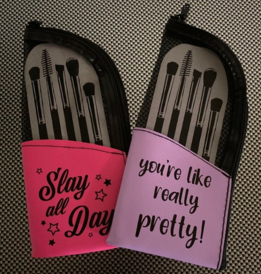 Two makeup brush sets in cases, one hot pink with "Slay all Day" and stars, the other light purple with "you're like really pretty!", each displaying five black brushes through a mesh top, resting on a black and white checkered surface