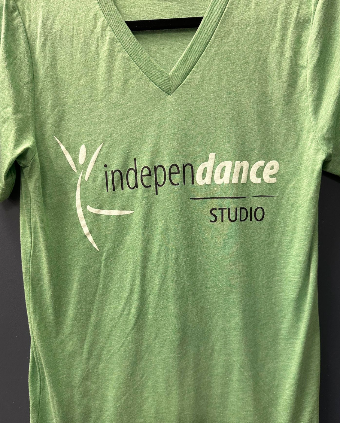 Photograph of a light green heathered V-neck t-shirt displaying a white stylized dancer logo next to the word independance in white text and STUDIO in black underlined text