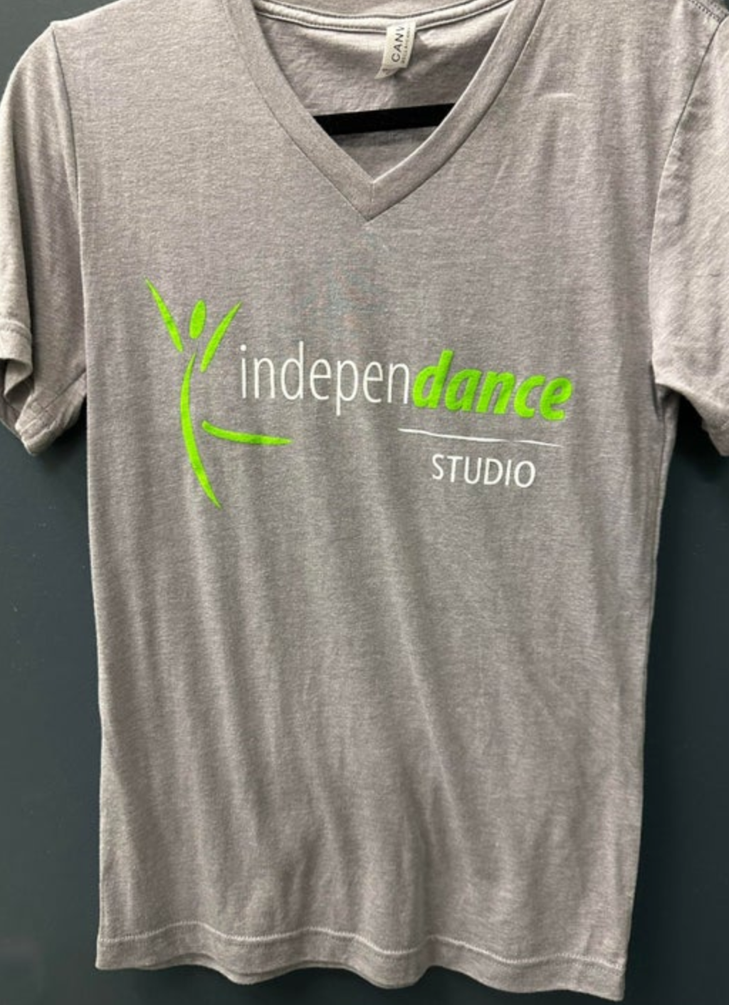 A gray v-neck t-shirt displaying the 'independance STUDIO' logo featuring a neon green abstract dancer and the word 'dance' highlighted in green, complemented by white hem stitching and a visible CANVAS tag