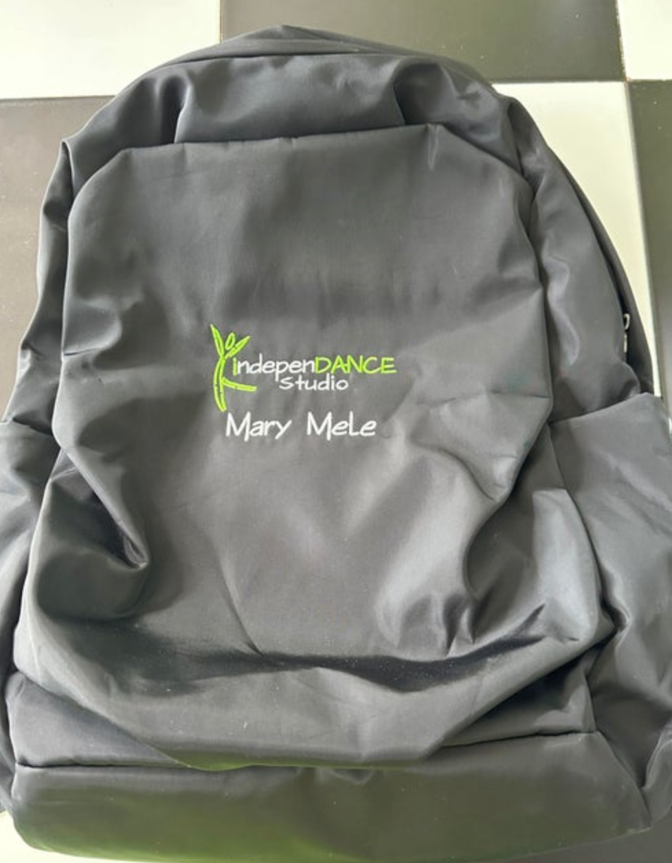 Photograph of a black backpack embroidered with a bright green indepenDANCE studio logo including a leaf design and white Mary Mele text, placed on a black and white checkered floor