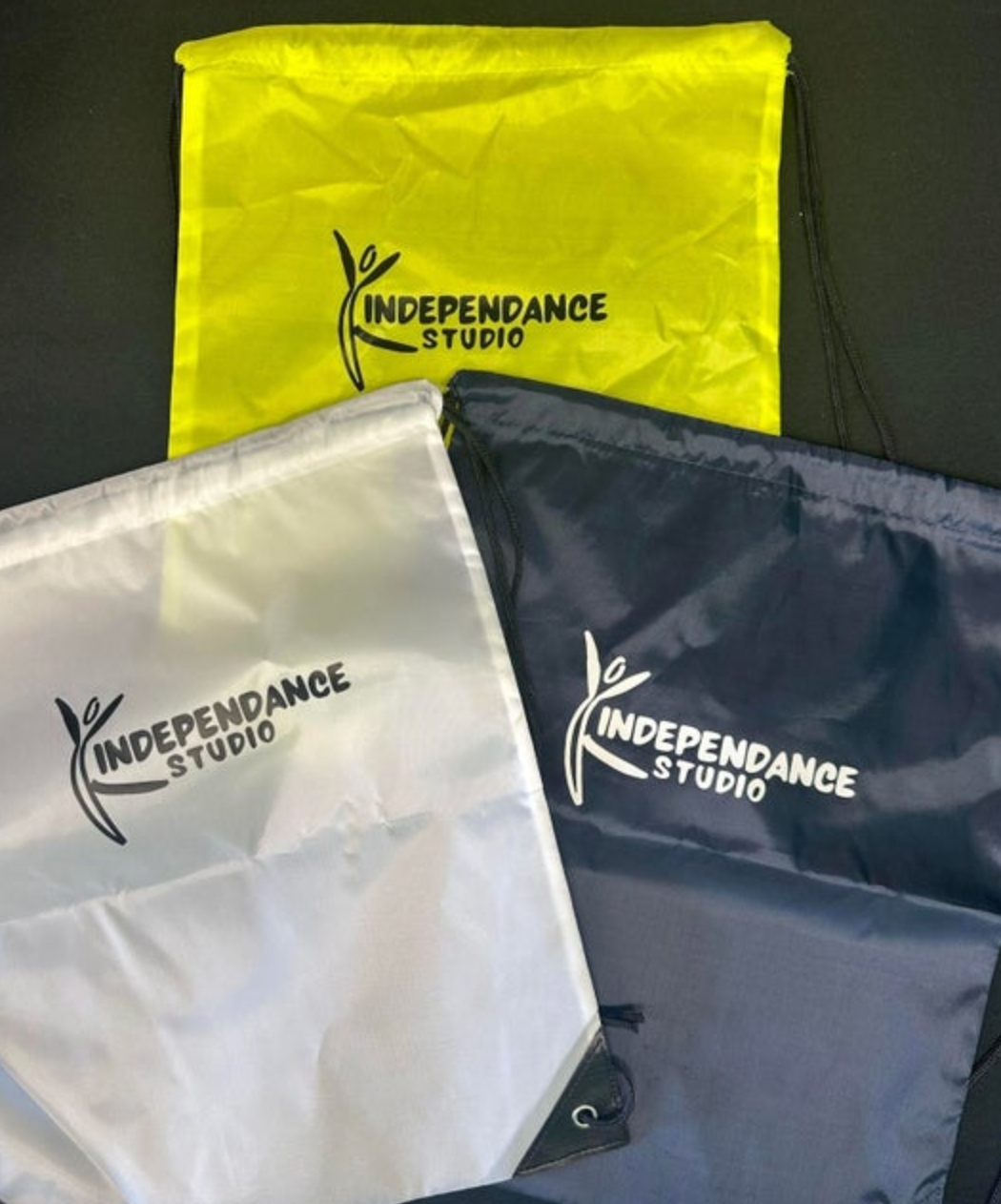 Three polyester drawstring bags in yellow, white, and navy blue, each displaying the black or white INDEPENDANCE STUDIO logo