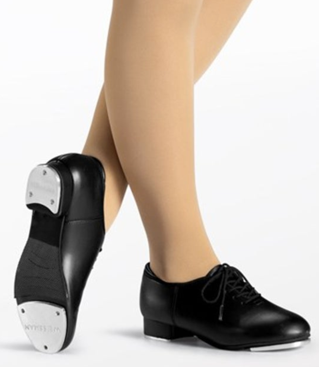 Photograph by Weissman of a dancer's crossed legs wearing black leather lace-up tap shoes with shiny silver toe and heel taps and nude dance tights on a white studio background
