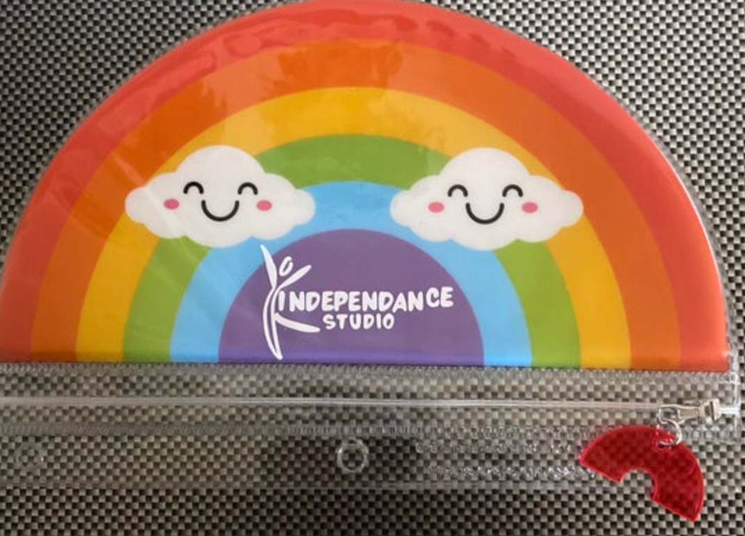 A vibrant rainbow arch pencil pouch featuring two smiling cartoon clouds and a white Independance Studio logo, with a clear zippered bottom half and a red rainbow charm zipper pull on a grey checkerboard background