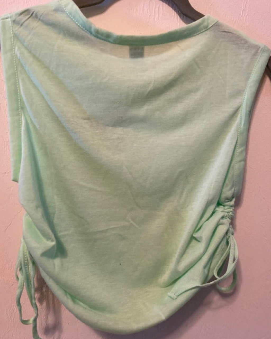 Light green sleeveless top hanging on a hanger against a pink wall featuring a round neck, gathered drawstring sides, and a subtly rounded hem