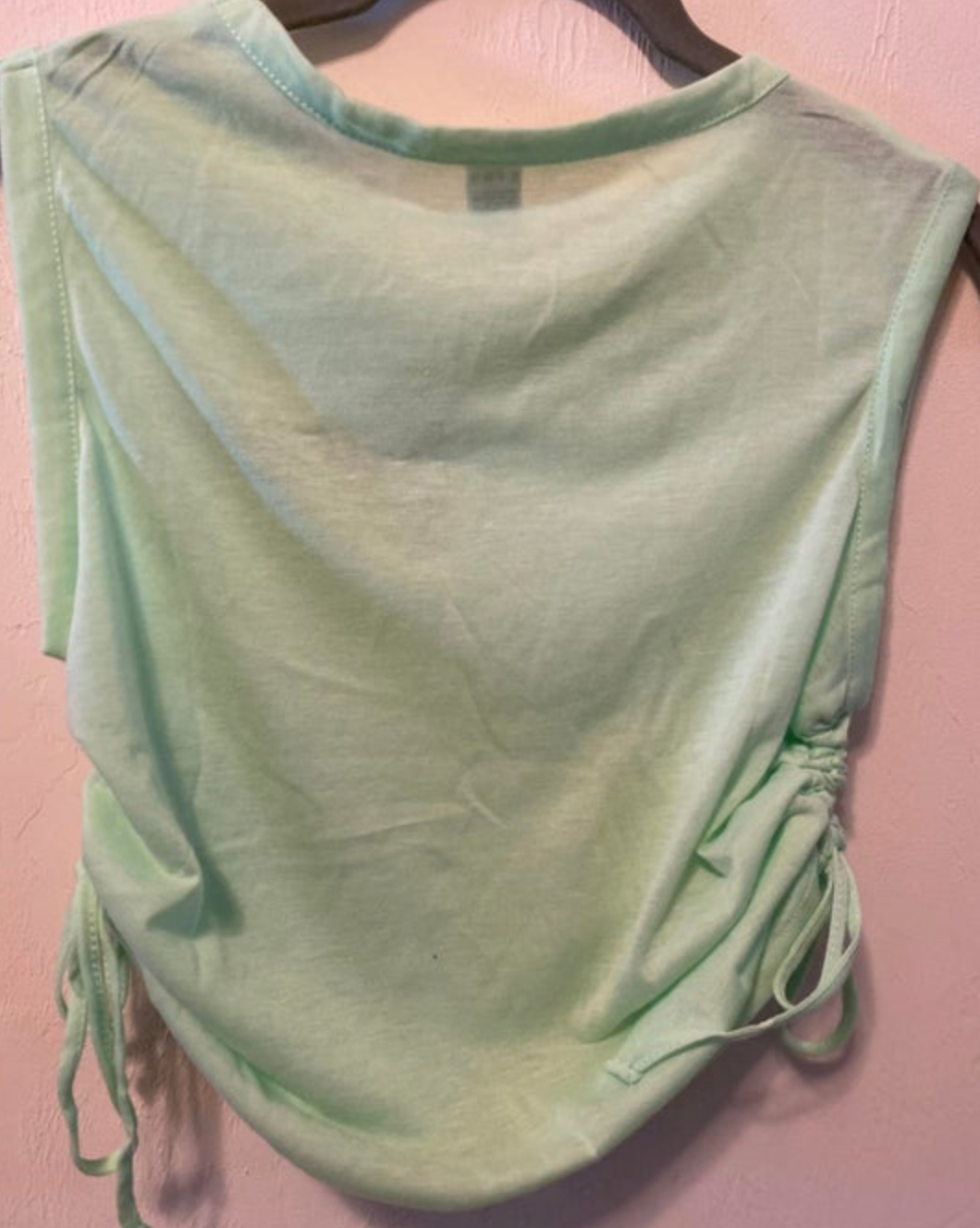 Light green sleeveless top hanging on a hanger against a pink wall featuring a round neck, gathered drawstring sides, and a subtly rounded hem