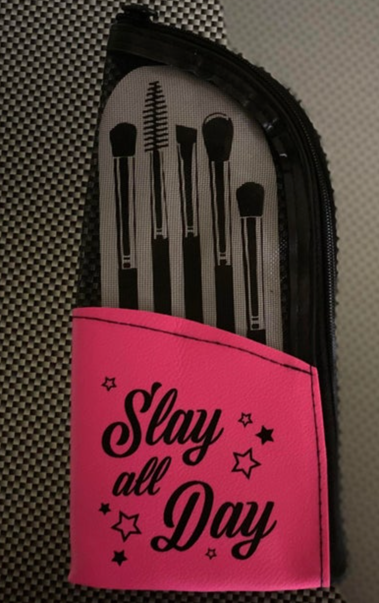 A black zippered makeup brush case featuring a vibrant hot pink "Slay all Day" flap with stars and a mesh panel revealing five black makeup brush illustrations resting on a checkered background