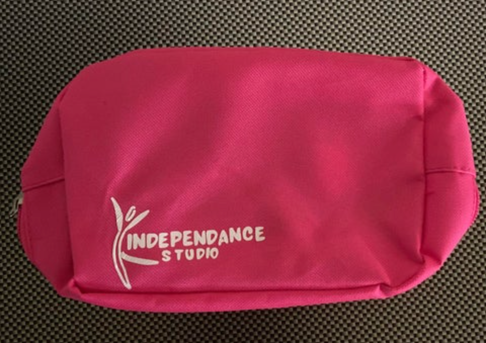 Bright pink fabric cosmetic bag featuring a white stylized dancing figure logo and the text INDEPENDANCE STUDIO resting on a grey checkered surface