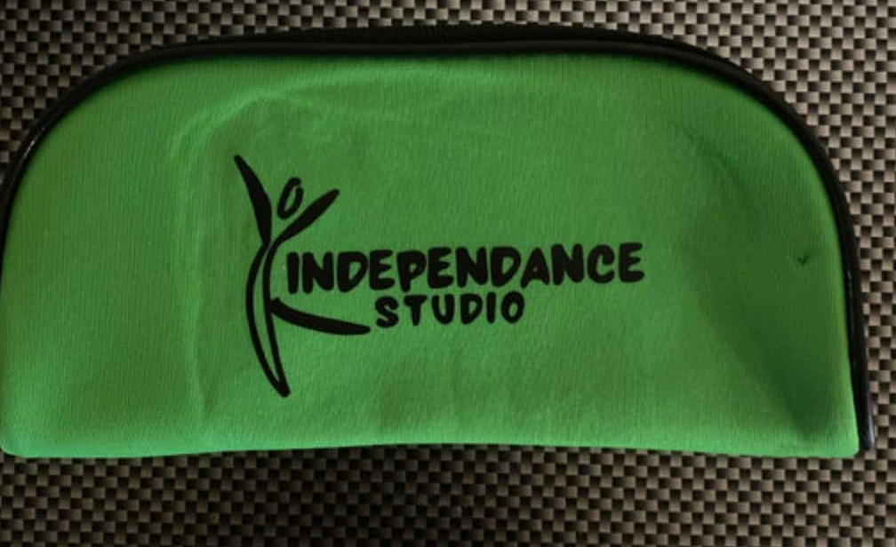 A fluorescent green textile pouch with a black stylized dancer logo and "INDEPENDANCE STUDIO" text, featuring a black trim, resting on a black and grey checkered surface
