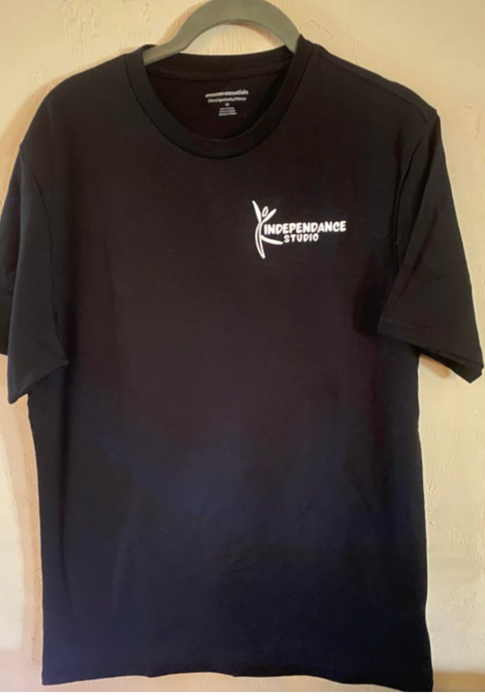 Black cotton t-shirt featuring a white INDEPENDANCE STUDIO logo with a stylized dancing figure, hanging on a grey plastic hanger against a textured light wall, revealing an Amazon Essentials neck label