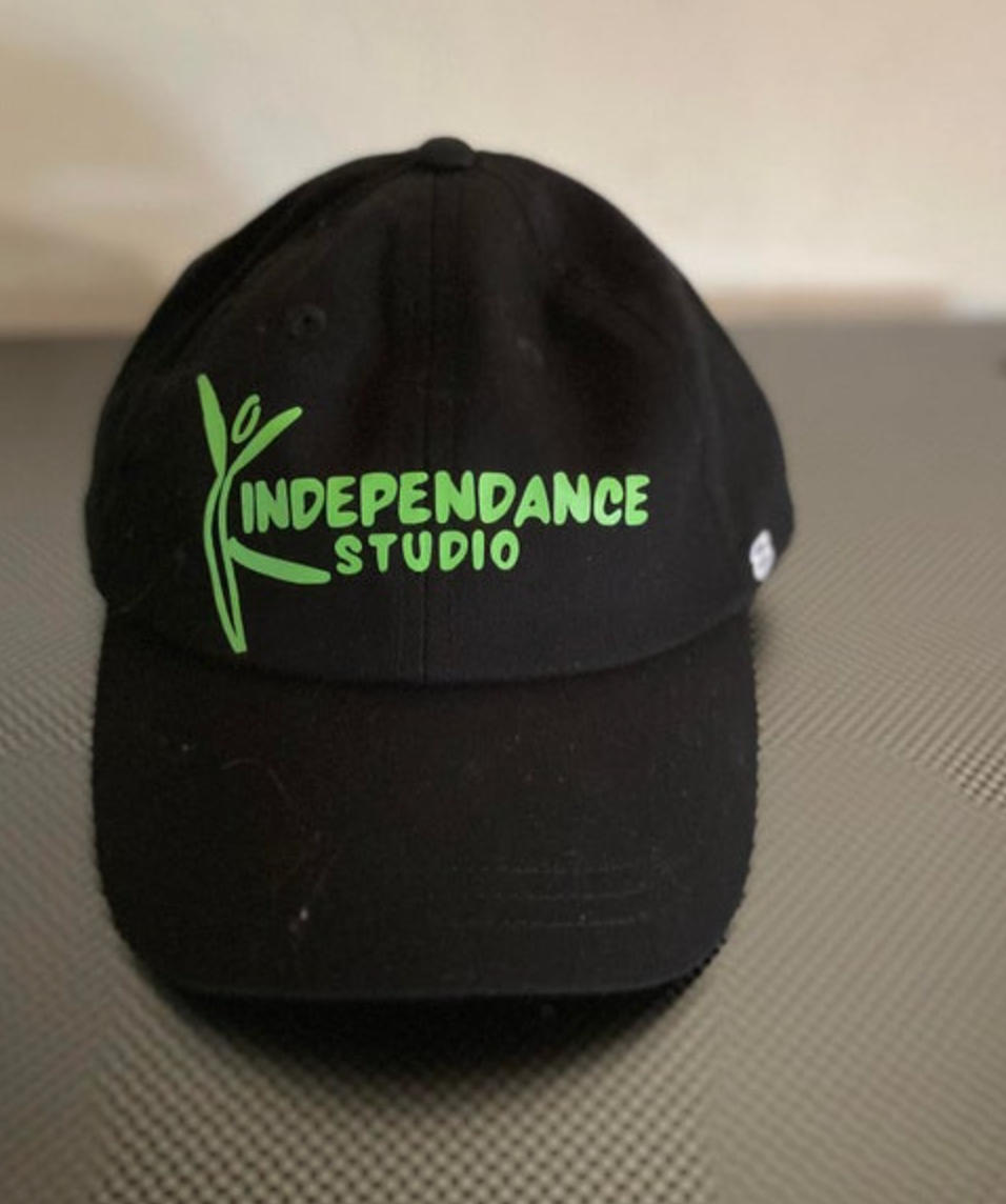 Photograph of a black baseball cap emblazoned with a vibrant green stylized dancer logo and INDEPENDANCE STUDIO text, resting on a dark patterned surface
