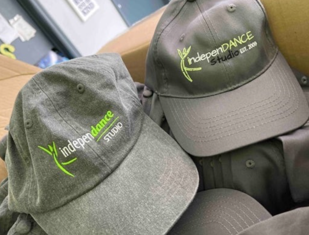 Product image of two charcoal grey baseball caps from IndepenDANCE Studio with a bright green logo, one displaying "indepenDANCE STUDIO" and the other "indepenDANCE Studio EST. 2009" piled in a box