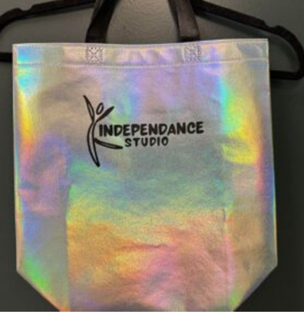 A holographic iridescent tote bag with a black Independance Studio dancer logo and black fabric handles