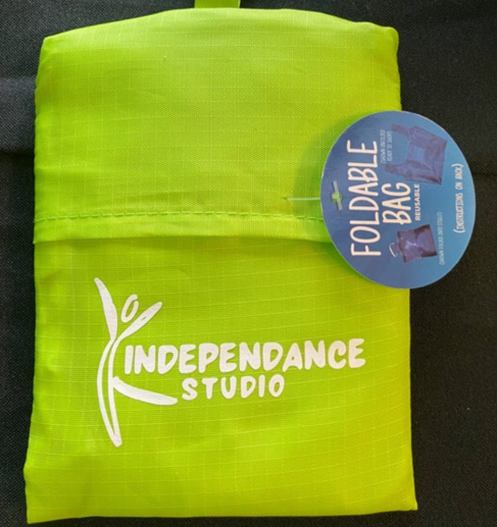 A fluorescent lime green Independance Studio foldable reusable bag features a prominent white logo and a blue circular tag illustrating its use
