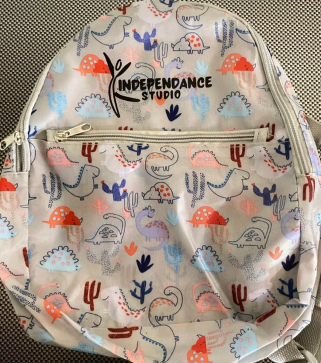 Light grey backpack with a playful pattern of colorful cartoon dinosaurs and stylized cacti alongside a black INDEPENDANCE STUDIO logo