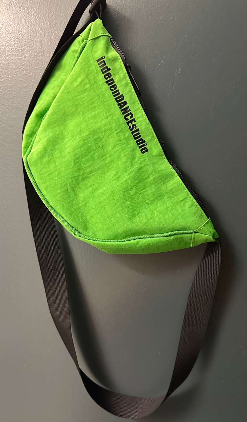 A bright neon green fanny pack with a visible black zipper and strap displaying the black text indepenDANCEstudio hanging on a gray wall