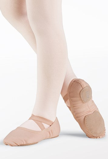 A dancer's feet and lower legs clad in light pink tights showcasing nude canvas ballet slippers featuring criss-cross elastic straps and visible split suede sole patches on a clean white studio background